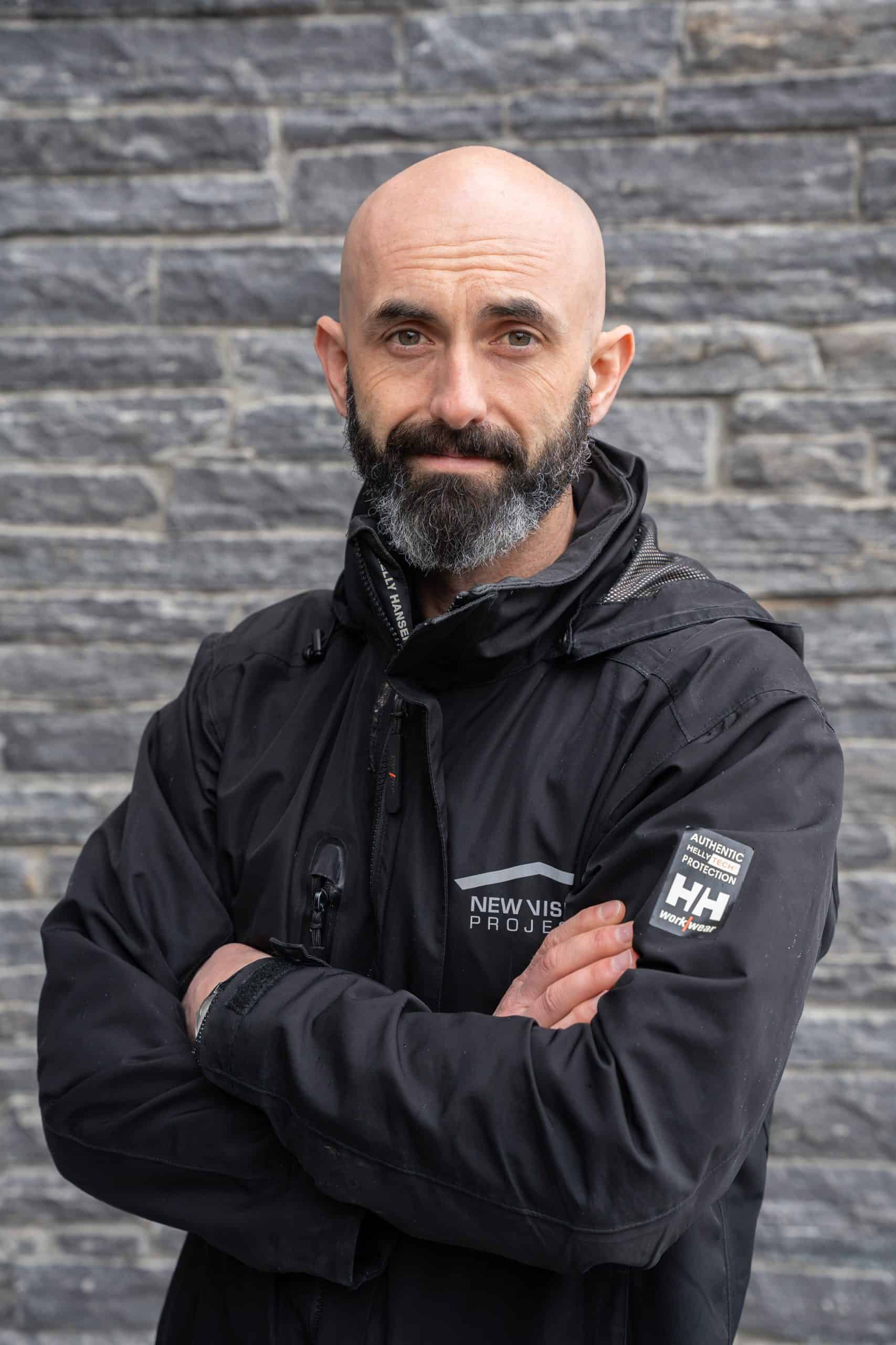 A confident man with a beard and bald head who is Brandon Smith of New Vision Projects, wearing a black jacket with branding, standing against a textured gray stone wall.